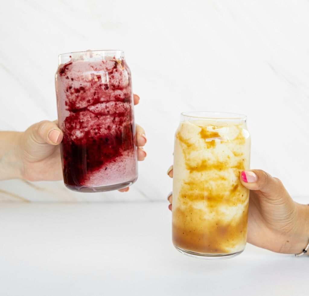 Two hands holding colorful iced drinks in clear glass jars against a bright backdrop.
